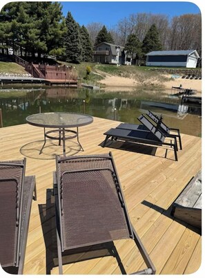 A wooden deck with a glass table and two lounge chairs, overlooking a calm body of water and a dock. - SilverLake House with Hot Tub, game room, huge yard, 1 mile from sand dunes. (Mears)