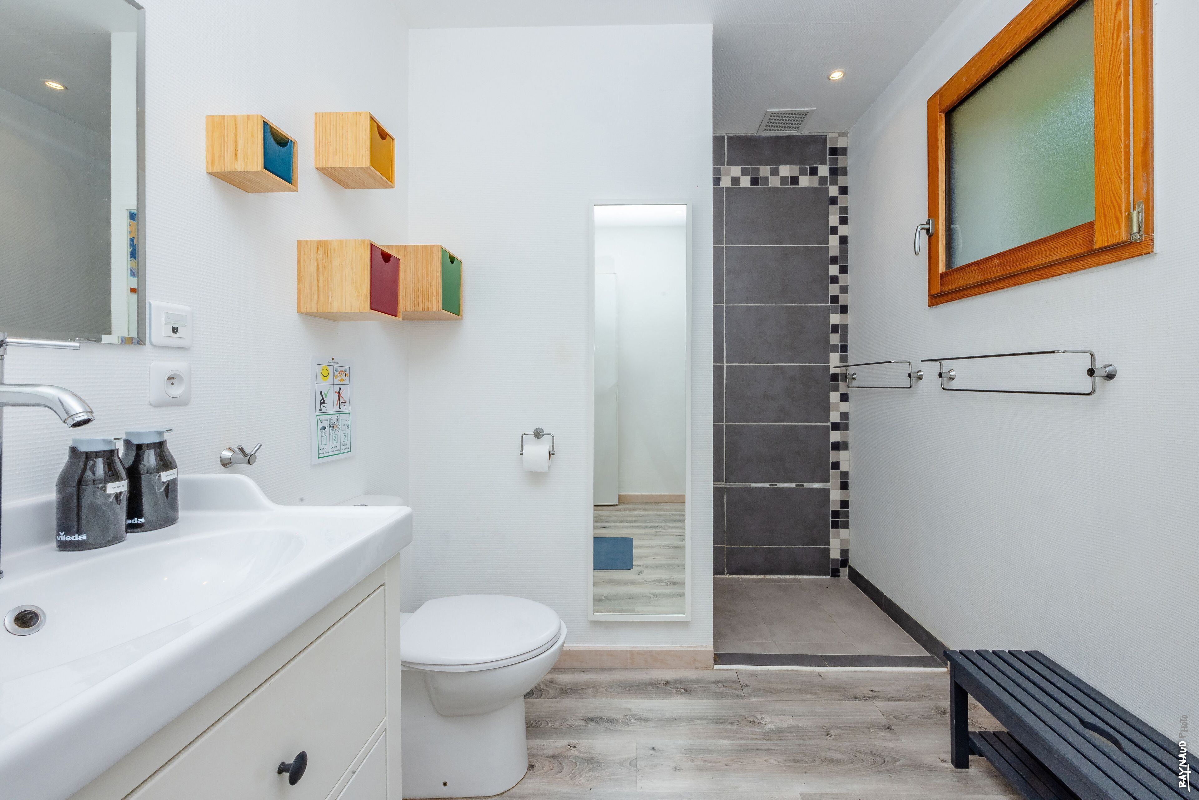 Ein modernes Badezimmer mit einem weißen Waschbecken, einer Toilette und einem Duschbereich. An der Wand befinden sich Holzaufbewahrungsboxen und eine Sitzbank im Duschbereich.