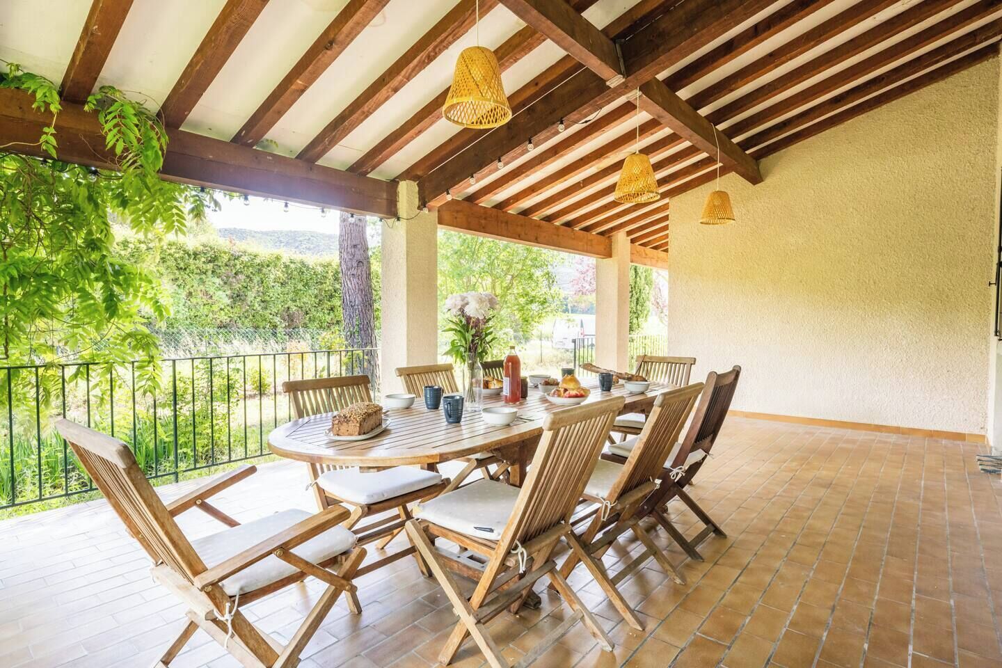 A covered patio with a wooden table and chairs, a vase with flowers, and a view of greenery.