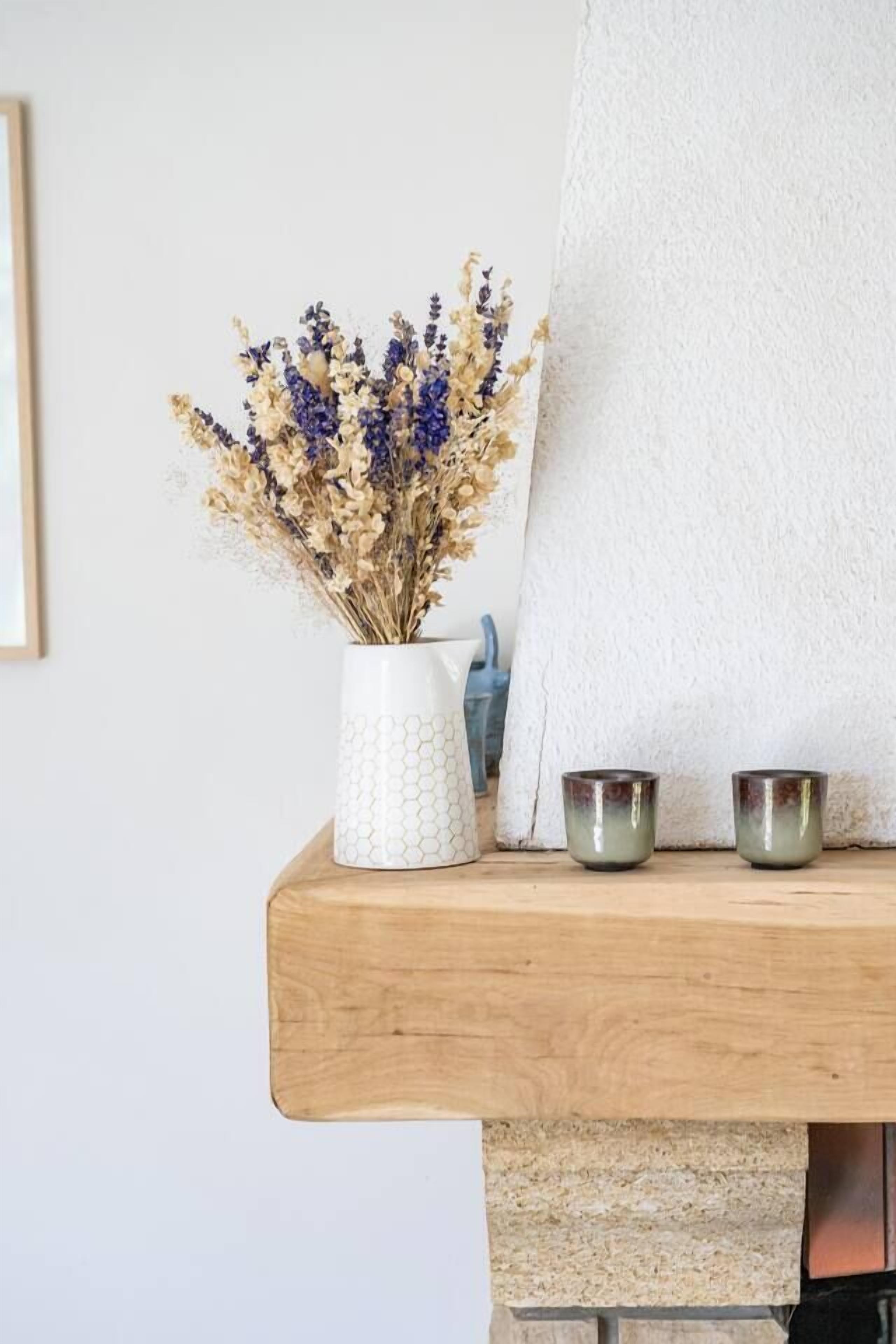 A white vase with dried flowers on a wooden shelf, a white towel, and two small metal cups.