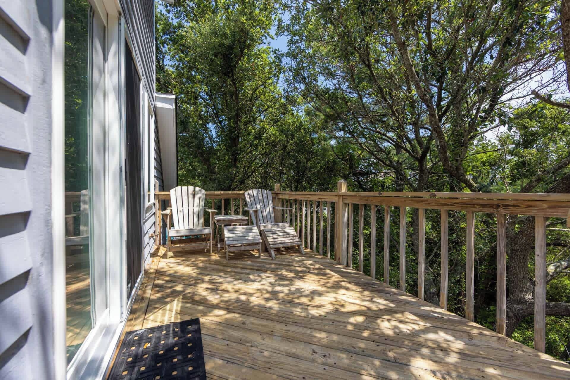 A wooden deck with Adirondack chairs and a wooden railing, overlooking a wooded area.