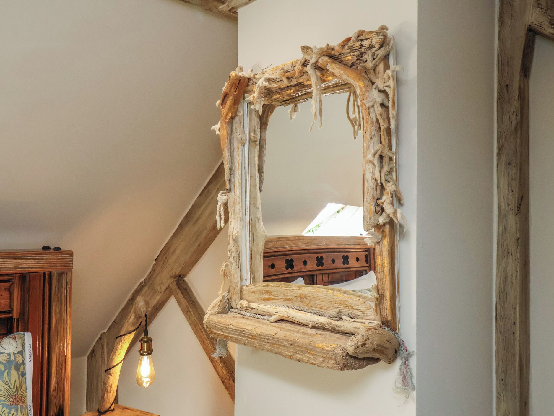 A rustic mirror with a wooden frame, mounted on a white wall in a room with exposed beams and a hanging light fixture.