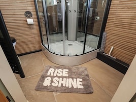 Zona de ducha con paredes de vidrio y una alfombra con la inscripción 'RISE & SHINE' en primer plano.