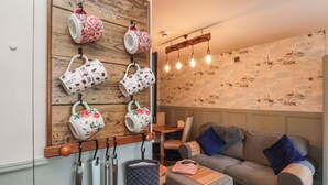 Ein gemütliches Wohnzimmer mit einem an der Wand befestigten Holzregal, das dekorative Tassen beherbergt, einem grauen Sofa mit blauen Kissen und einer Wand mit Muster-Tapete.