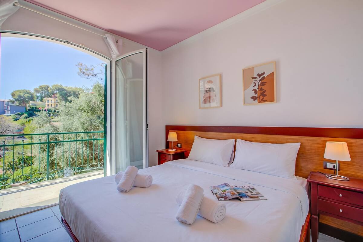 A hotel room with a bed, two bedside tables with lamps, a balcony with a view, and two framed pictures on the wall.