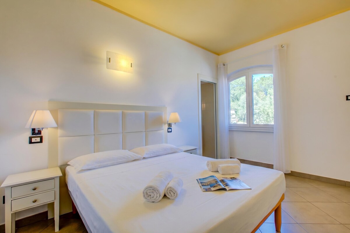 A hotel room with a bed, nightstands, lamps, towels, and a window with curtains.