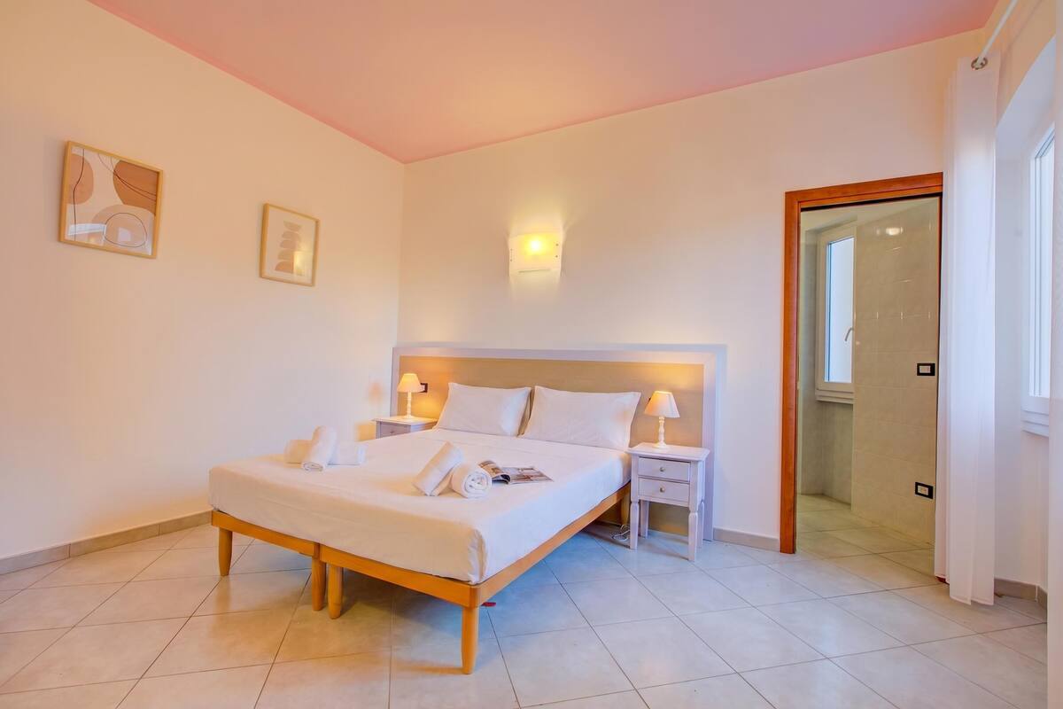 A bedroom with a bed, bedside tables, a nightstand, a wall-mounted light, and a door leading to a bathroom.