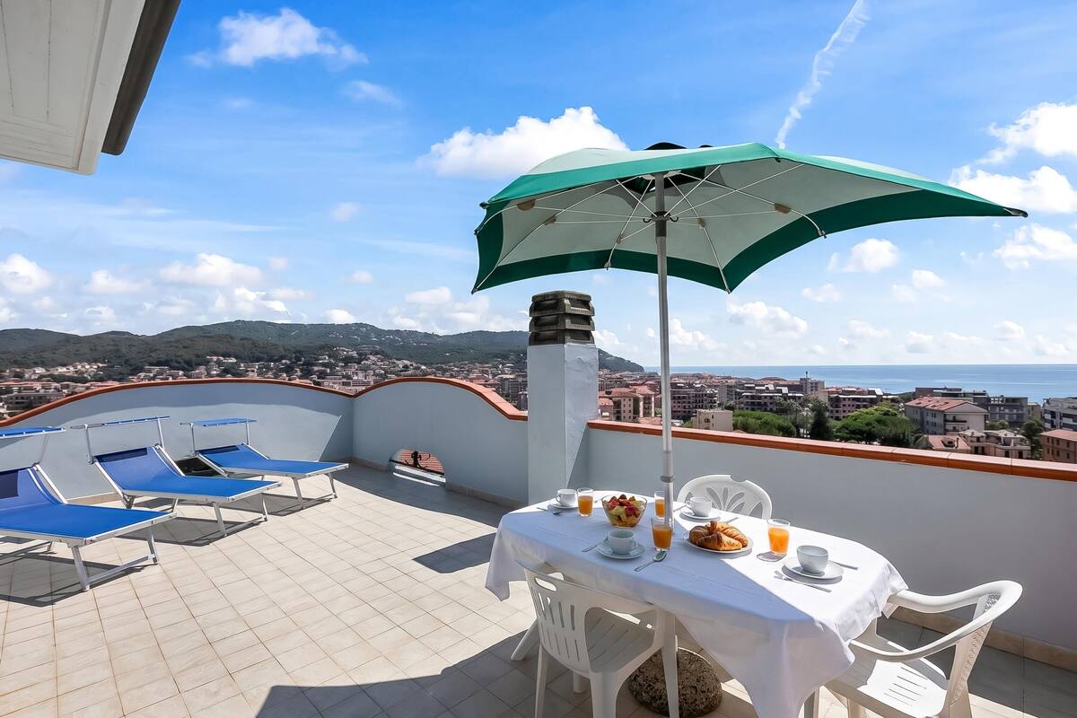A terrace with a table set for breakfast, blue lounge chairs, and a green umbrella.