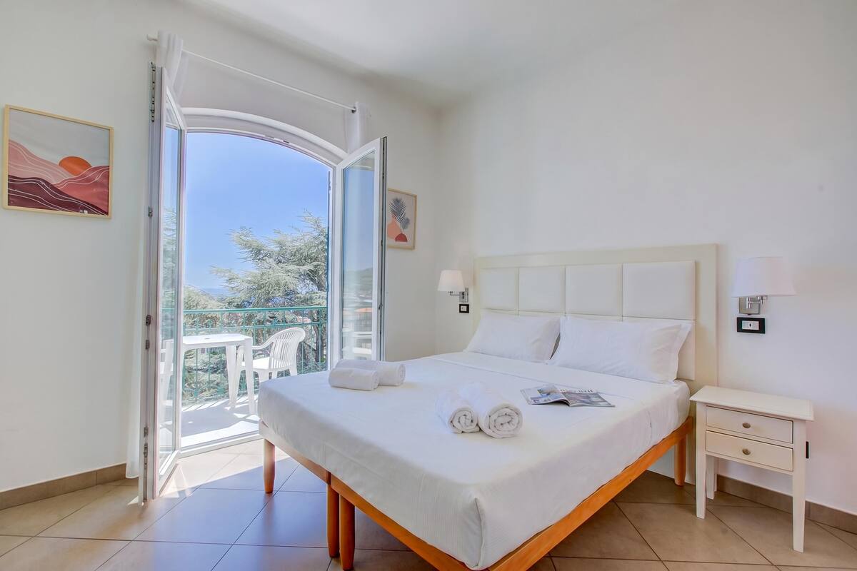 A hotel room with a bed, a nightstand, a window with curtains, and a balcony with chairs.