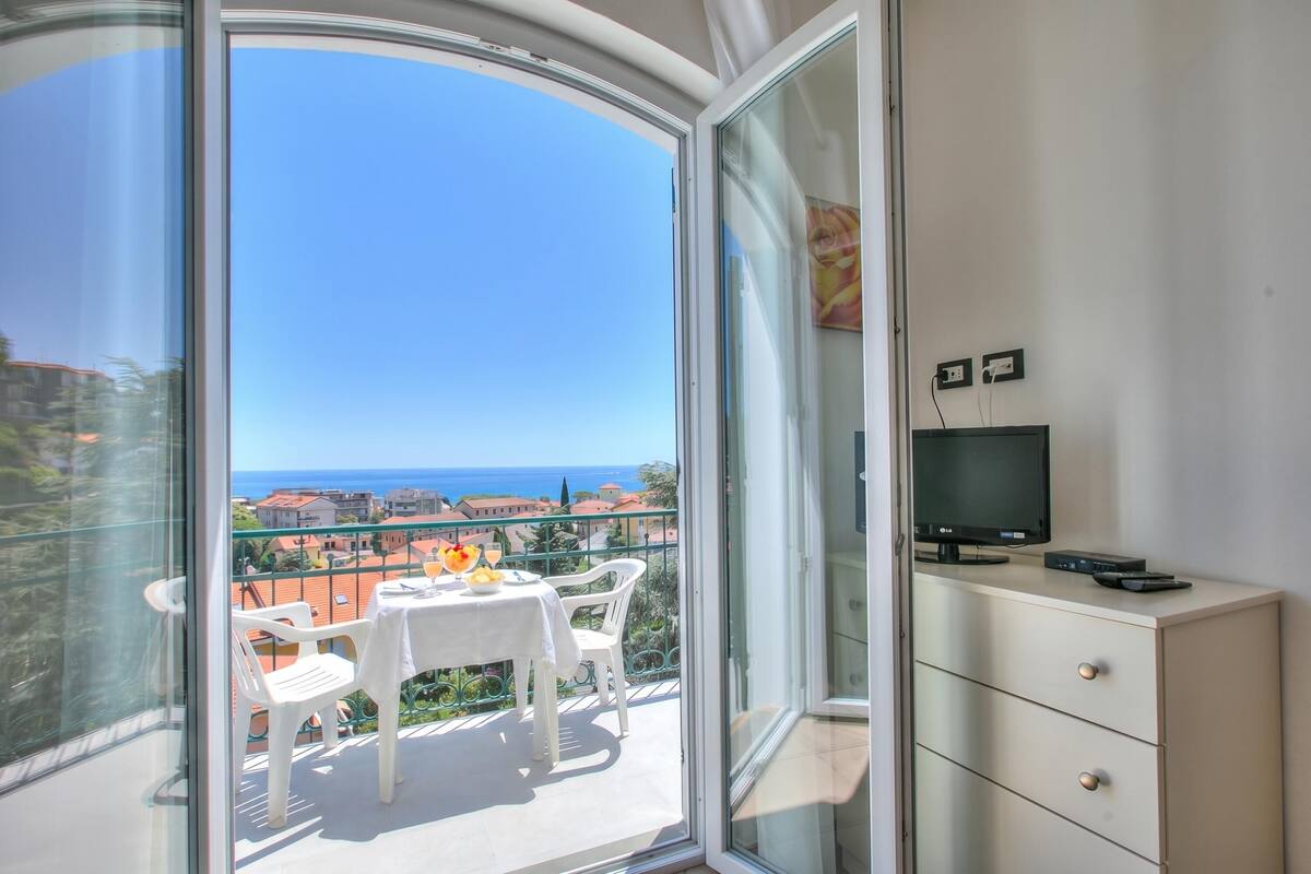 A balcony with a table and chairs, a TV on a cabinet, and a clear view of the sea and buildings.