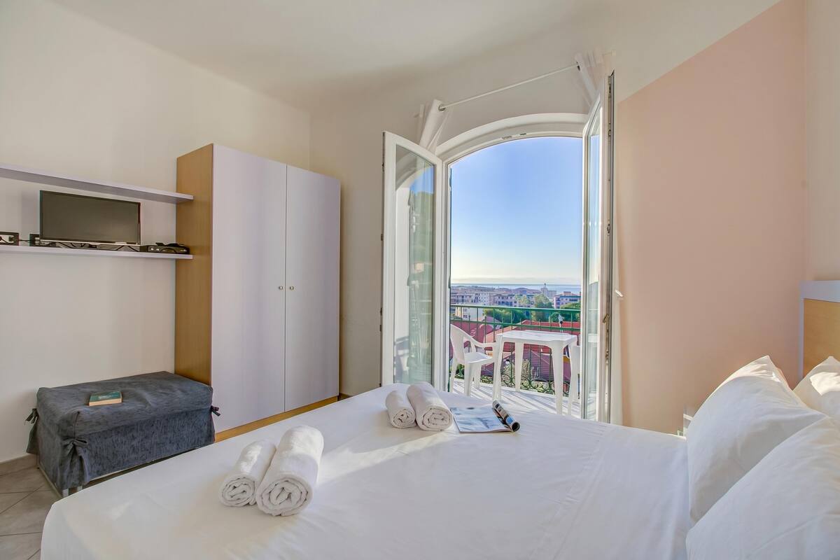 A hotel room with a bed, a desk, a wardrobe, and a balcony with a city view.