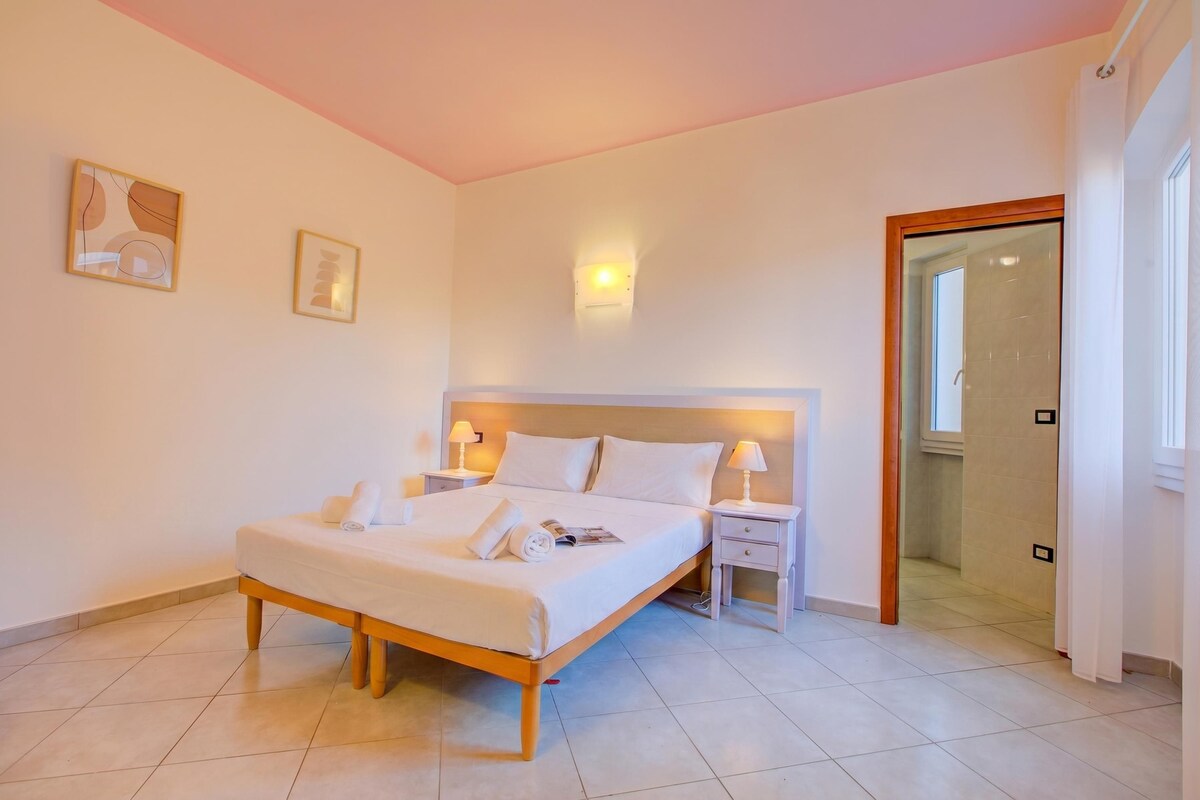 A bedroom with a bed, bedside tables, a nightstand, a wall-mounted light, and a door leading to a bathroom.