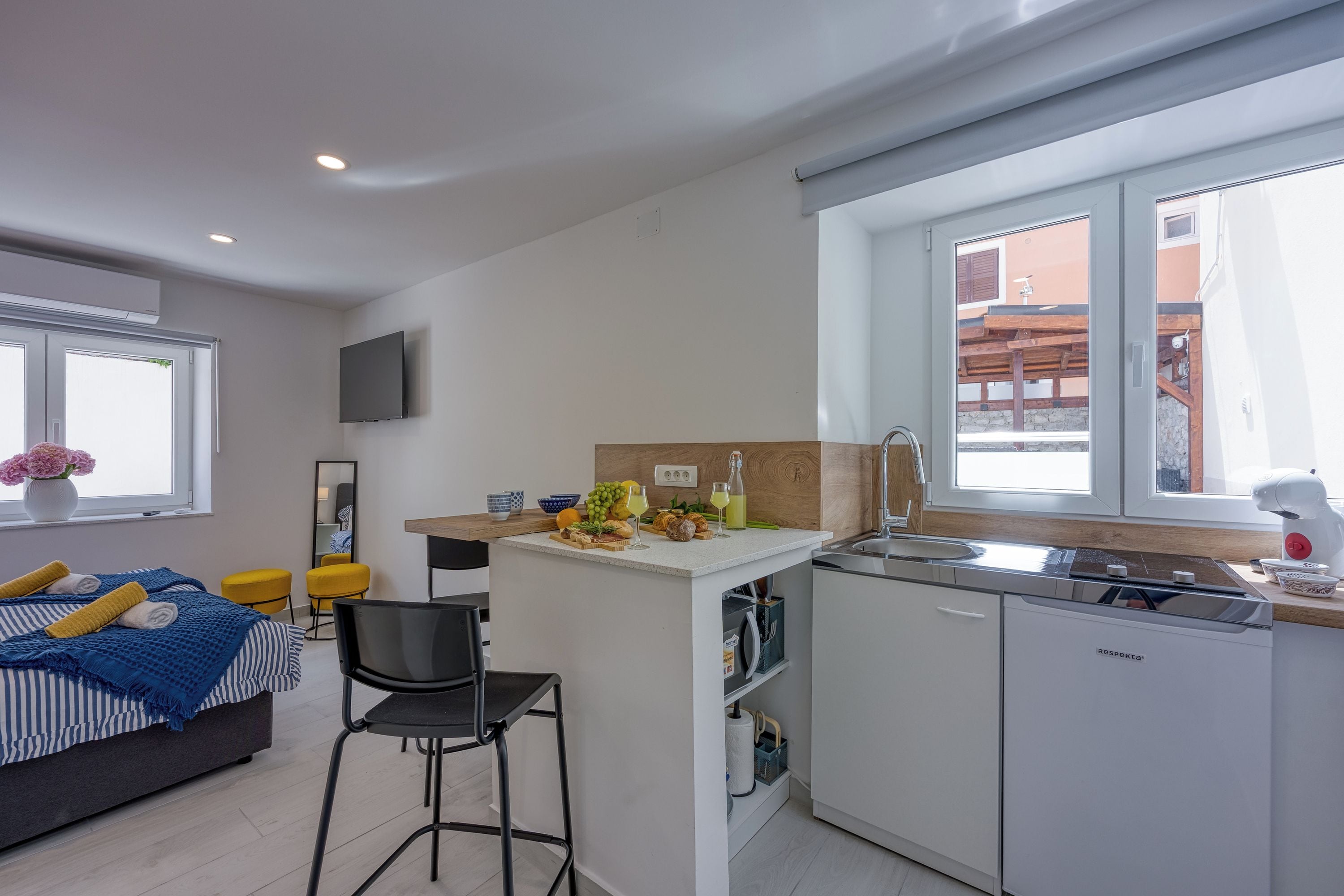 A modern kitchen with a central island, a dining area with a chair, a bed with a blue and white striped bedspread, and a large window with a view of buildings.