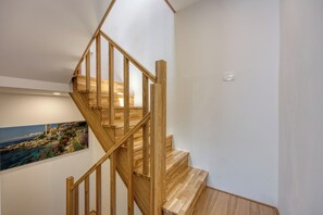 Un escalier en bois avec une rampe, un interrupteur fixé au mur et un tableau représentant un paysage côtier.