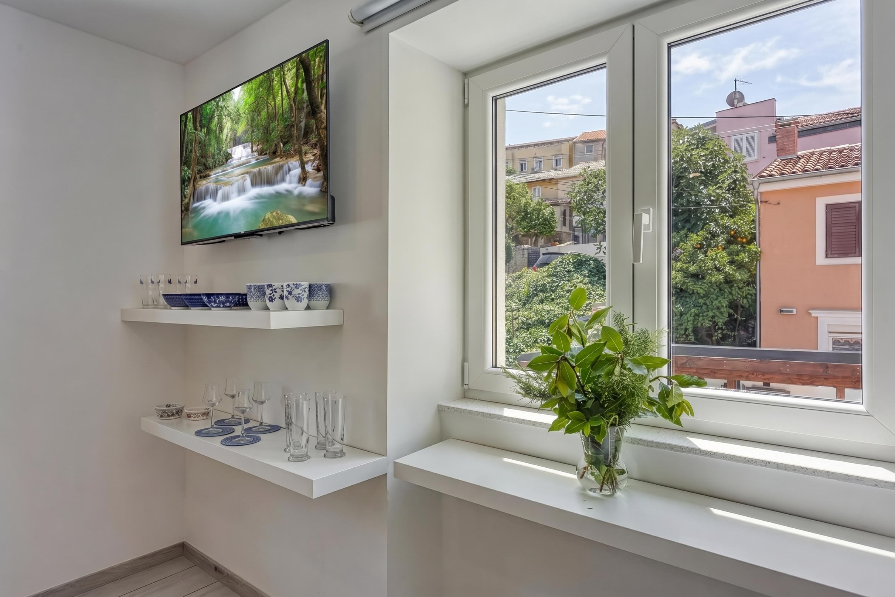 A modern room with a TV on the wall, a window with a view of buildings, and a shelf with glassware.
