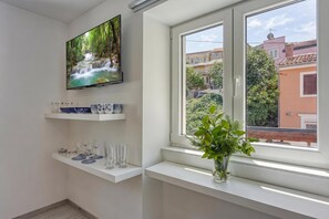 Una habitación moderna con un televisor en la pared, una ventana con vistas a edificios y un estante con cristalería.
