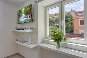 A modern room with a TV on the wall, a window with a view of buildings, and a shelf with glassware.