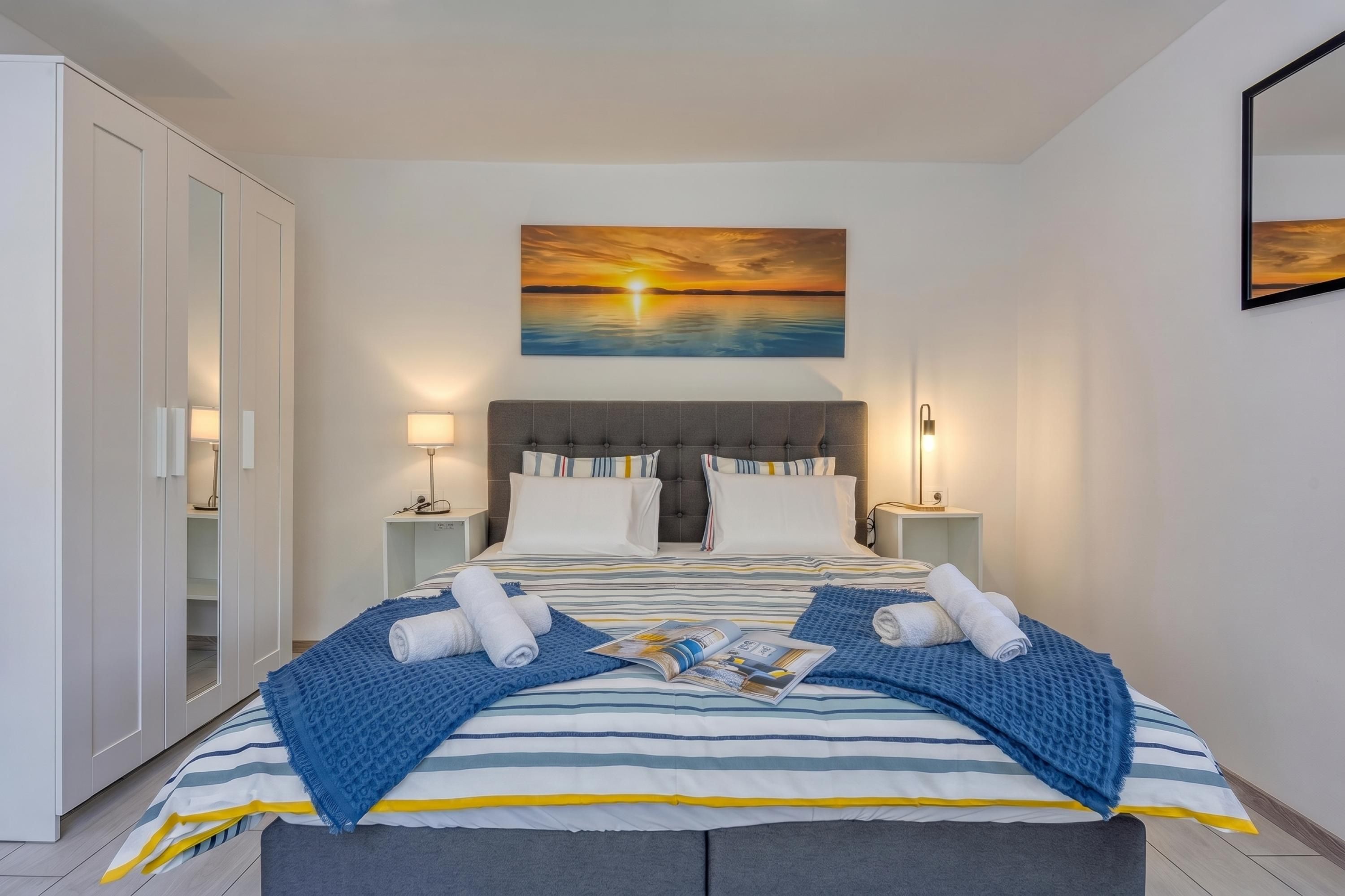 Cama bem arrumada com roupa de cama listrada, duas luminárias de cabeceira, um quadro com um pôr do sol e um cobertor azul.
