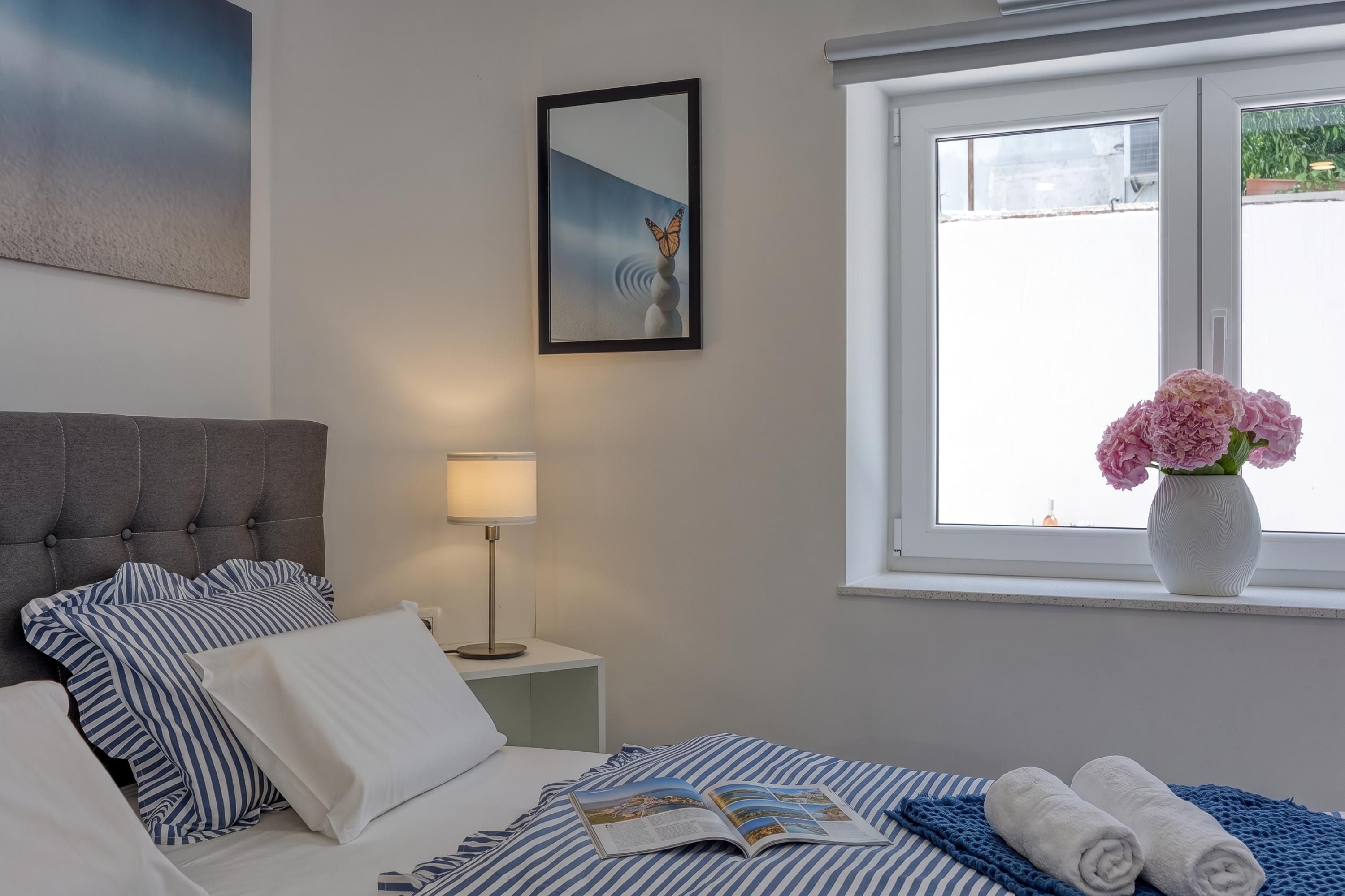 A hotel room with a bed, a bedside table with a lamp, a framed picture on the wall, and a window with a view of the outside.