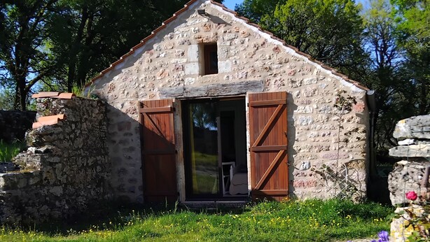 Un edificio rústico de piedra con techo inclinado y contraventanas de madera, rodeado de follaje y flores.