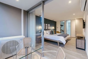 A modern bedroom with a glass partition, a bed, a chair, a small table, and a wall-mounted TV.