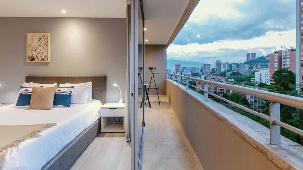 Un dormitorio moderno con una cama grande, mesita de noche y vistas a un paisaje urbano a través de la barandilla de un balcón.