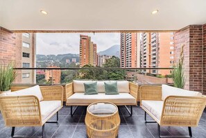 A balcony with wicker furniture, a sofa with cushions, and a view of tall buildings and greenery.