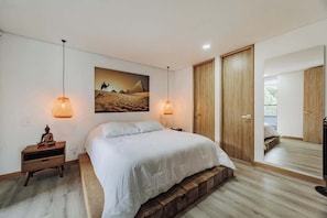 A bedroom with a bed, wooden floor, two pendant lights, a nightstand, a wall-mounted painting, and a mirror.