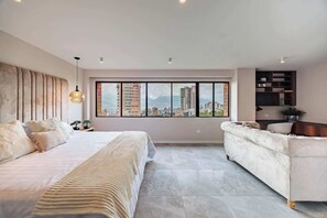 Un dormitorio moderno con una cama grande, un sofá, vistas a la ciudad a través de amplios ventanales y una estantería empotrada.