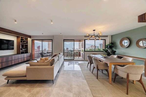 Una sala moderna con sofá, mesa de comedor y sillas, ventanas amplias con vista a la ciudad y un televisor de pantalla plana montado en la pared.