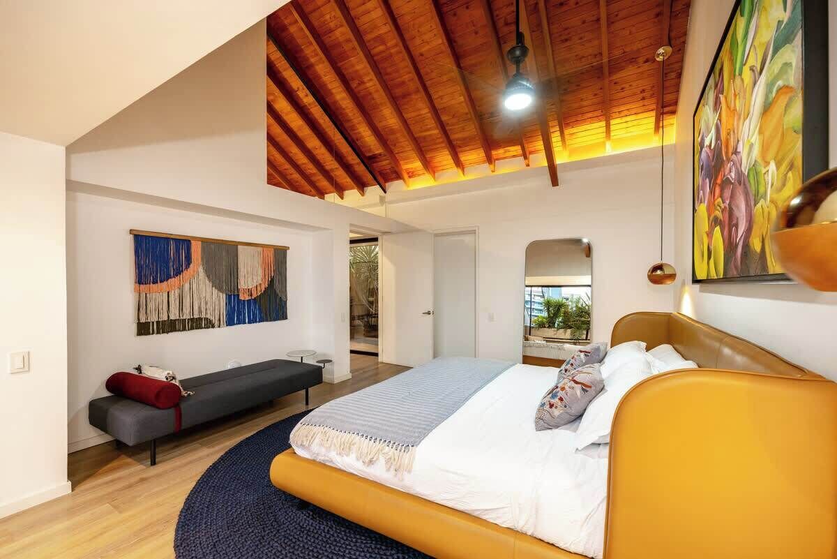 A bedroom with a large bed, a bench, a painting, and a ceiling fan.