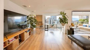 A modern living room with a large flat-screen TV, wooden flooring, a black leather bench, and a dining area with a table and chairs. - |EP| Premium PH W/Sauna, Jacuzzi, SteamRoom (Medellín)