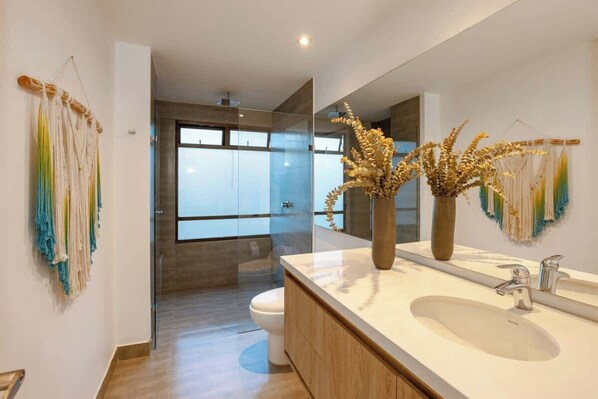 A modern bathroom with a large mirror, two sinks, and a toilet. There are decorative plants and wall art.