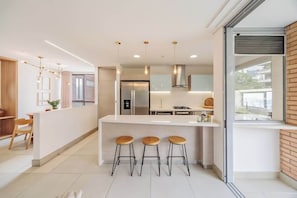 A modern kitchen with a central island, bar stools, pendant lighting, and a view of the outside.