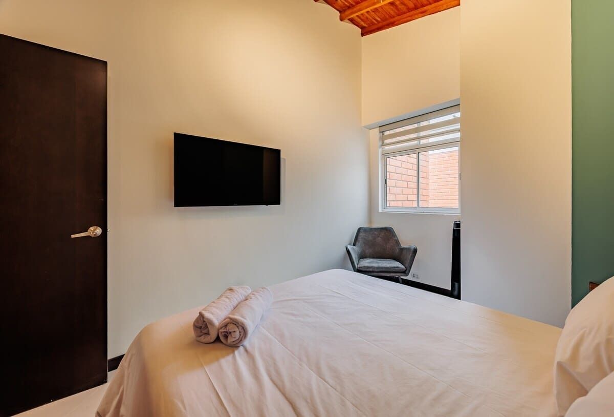 A hotel room with a bed, a television mounted on the wall, a window with blinds, and a chair.