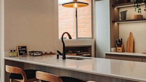 Una cocina moderna con una isla central, taburetes de bar, una lámpara colgante y una pared de ladrillo visible a través de la ventana.