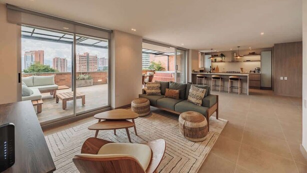 Amplia sala de estar con sofá, mesa de centro y reposapiés, una cocina con taburetes de bar y ventanas grandes que ofrecen vista a la ciudad.