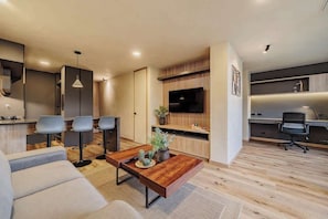 Una sala moderna con un sofá, una mesita de centro y un televisor. Cuenta con una zona de cocina con taburetes altos y una mesa de comedor.