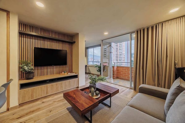 Una sala moderna con un televisor de pantalla plana montado en una pared de paneles de madera, una mesa de centro de madera con objetos decorativos, un sofá y un ventanal corredero que da a un balcón con vistas a la ciudad.