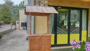 A wooden structure with a slanted roof, possibly a shelter or kiosk, in front of a building with a yellow awning and glass doors. - Hotel Criss (Rimini)