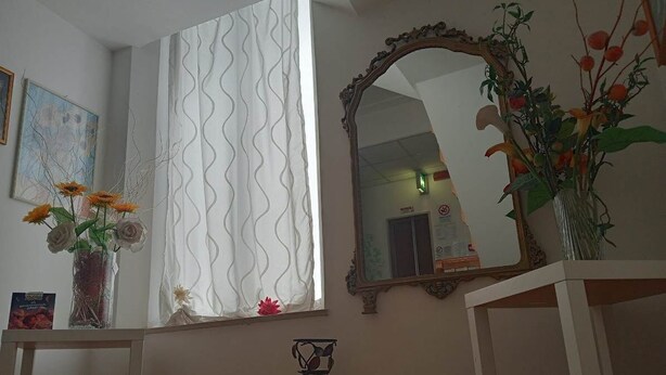 A room with a large window covered by a wavy curtain, a mirror with an ornate frame, and two tables with vases of flowers. - Hotel Criss (Rimini)