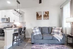 Eine moderne Küche mit weißen Schränken, einer eingebauten Mikrowelle und einer Bar mit Hockern. Im Wohnbereich befindet sich ein graues Sofa mit Karusschalldekorationen sowie zwei gerahmte Bilder an der Wand.
