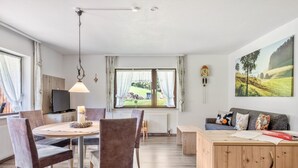 Una sala moderna con comedor, un sofá, un mueble de madera y vistas a un paisaje verde por la ventana.