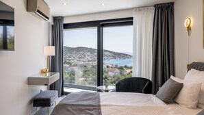 Une chambre d’hôtel moderne avec un grand lit, une table de chevet, une chaise et une vue sur une ville côtière.