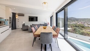A modern dining area with a wooden table and grey upholstered chairs, a kitchen with white cabinets, and a large window offering a view of a residential area. - Mar Escape Designer Villa Croatia w/Sea Views, Private Pool, 4 En-Suite Bedrooms (Vinišće)