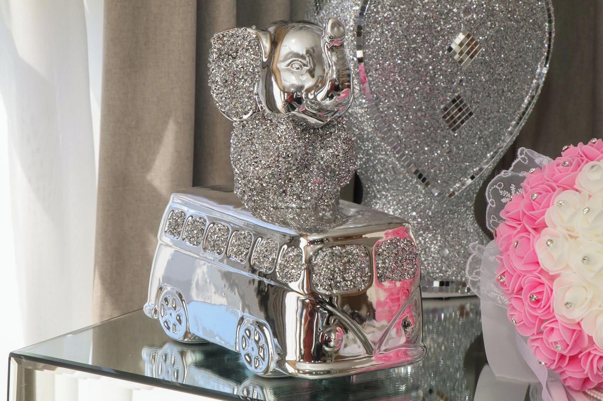 A silver decorative item with a face, a clock, and a vase of pink flowers on a glass table.