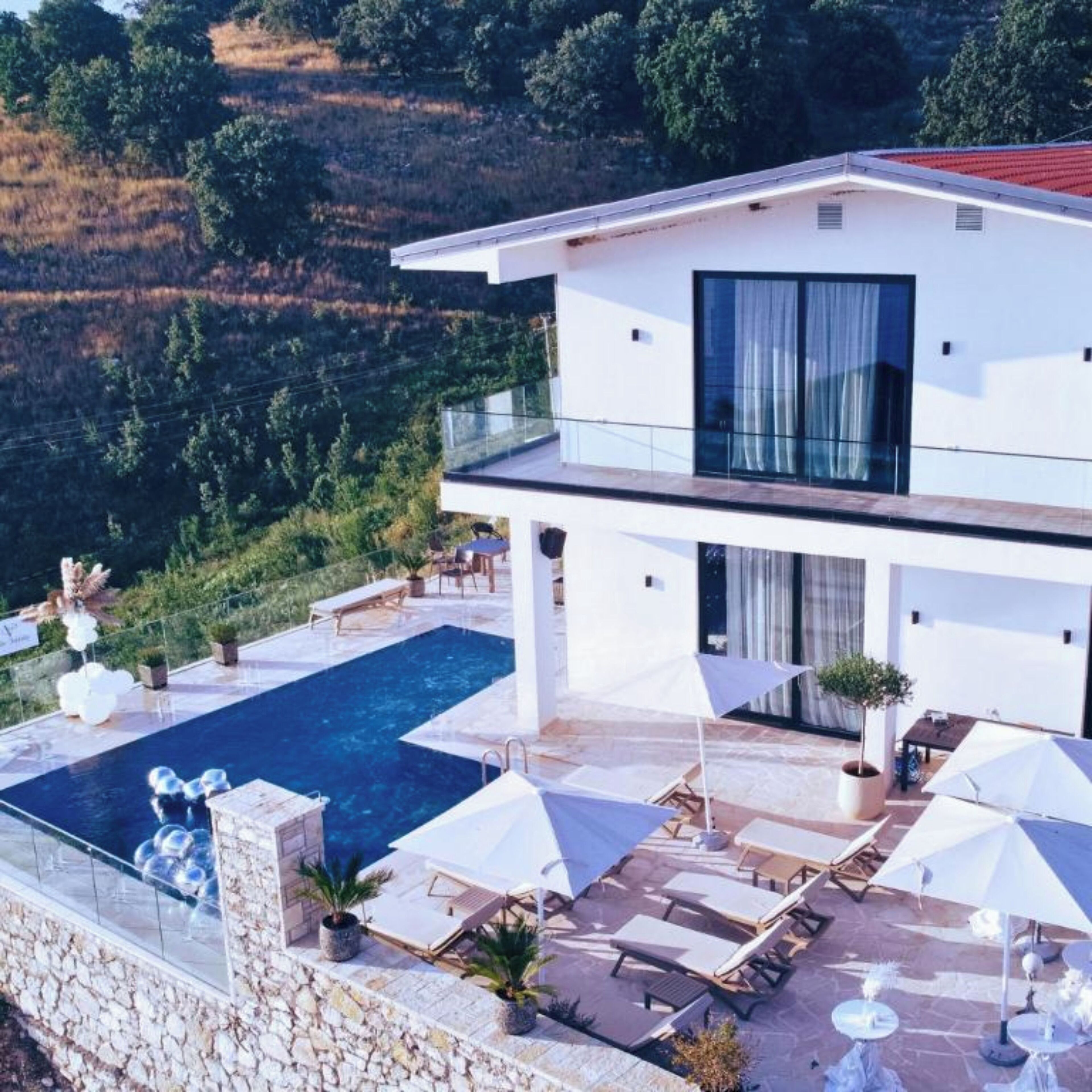 A modern two-story house with a large swimming pool on the ground floor, outdoor seating, and a view of the surrounding hills.