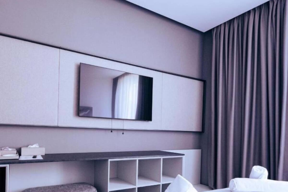 A modern hotel room with a large bed, a desk with a flat-screen TV, a grey ottoman, and a built-in shelving unit.