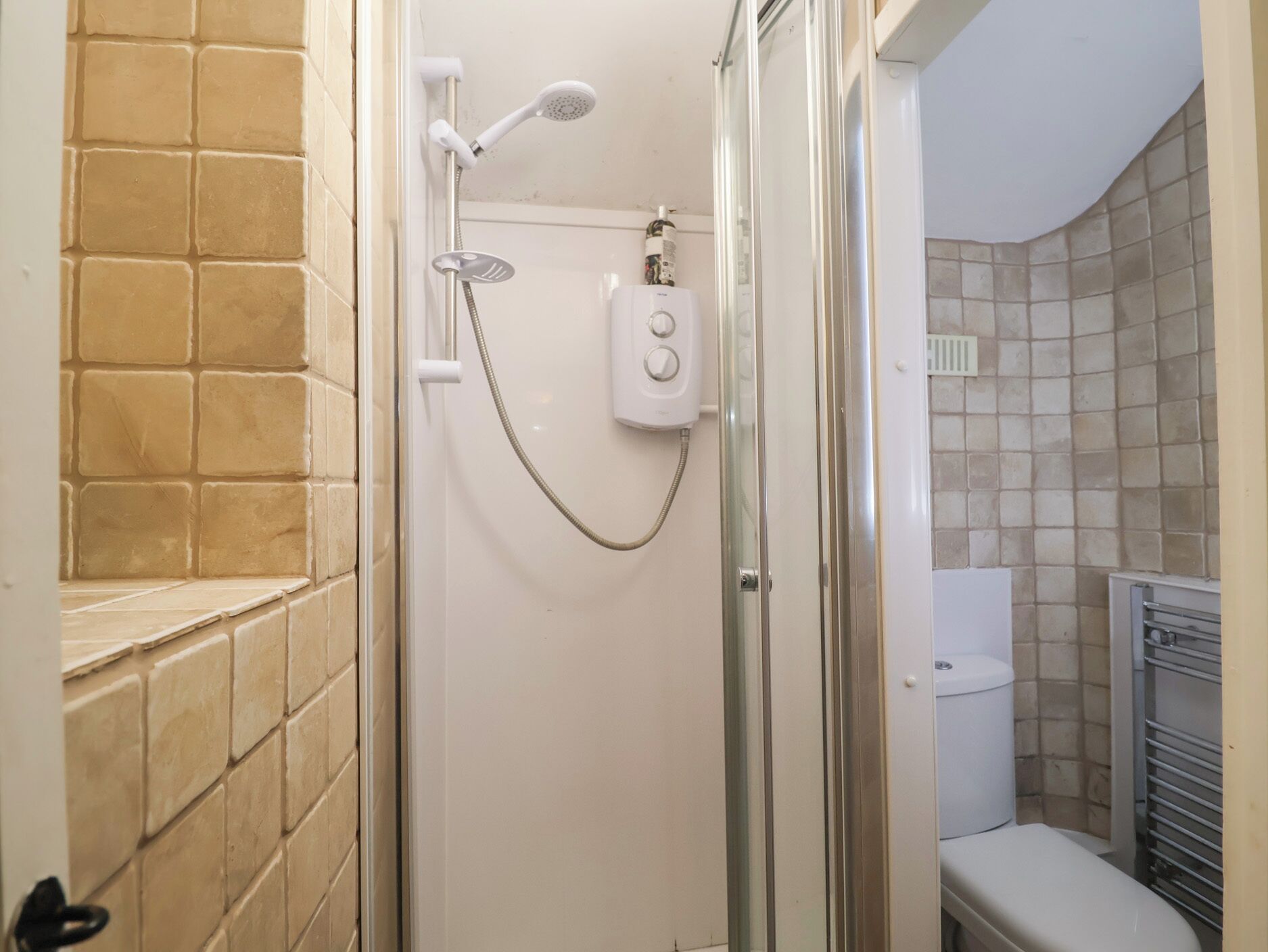 Une salle de bain avec une douche à parois de verre, des toilettes blanches et des murs recouverts de carreaux beiges.