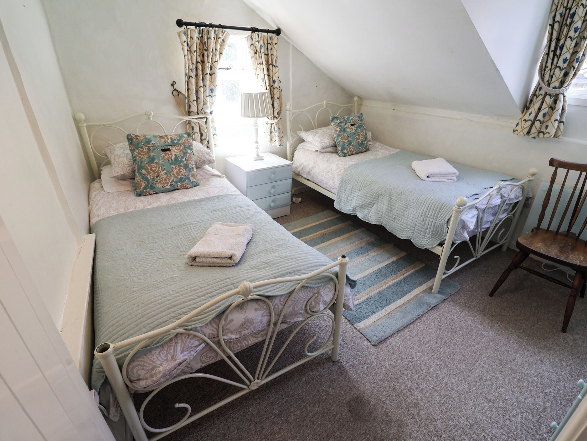 A cozy attic bedroom with two twin beds, a small bedside table, a chair, and a window with curtains.